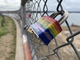 A rainbow-colored padlock is attached to a chain-link fence, with out-of-focus surroundings including a body of water and grassy ground. The padlock features the branding 'Master' on its blue base.