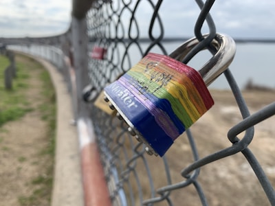 A rainbow-colored padlock is attached to a chain-link fence, with out-of-focus surroundings including a body of water and grassy ground. The padlock features the branding 'Master' on its blue base.
