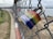 A rainbow-colored padlock is attached to a chain-link fence, with out-of-focus surroundings including a body of water and grassy ground. The padlock features the branding 'Master' on its blue base.
