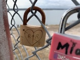 A close-up of a rusted padlock attached to a wire fence. The padlock has a heart with 'R + R' carved inside it, along with an arrow through the heart, symbolizing love. The background is slightly out of focus, showing a body of water and a distant landscape.