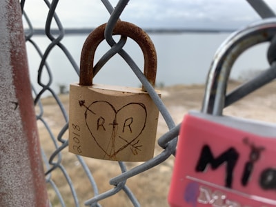 A close-up of a rusted padlock attached to a wire fence. The padlock has a heart with 'R + R' carved inside it, along with an arrow through the heart, symbolizing love. The background is slightly out of focus, showing a body of water and a distant landscape.