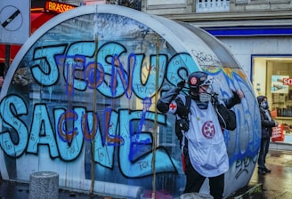 A person wearing protective gear, including a helmet and mask, stands in front of a large glass structure covered in colorful graffiti. The graffiti features bold letters in blue and purple. The scene takes place in an urban setting, with a shop visible in the background.