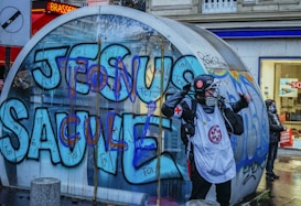 A person wearing protective gear, including a helmet and mask, stands in front of a large glass structure covered in colorful graffiti. The graffiti features bold letters in blue and purple. The scene takes place in an urban setting, with a shop visible in the background.