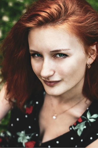 A close-up portrait of a person with auburn hair and bright green eyes, wearing a black dress with floral and polka dot patterns. The background appears to be a soft focus of greenery, enhancing the vividness of the image.