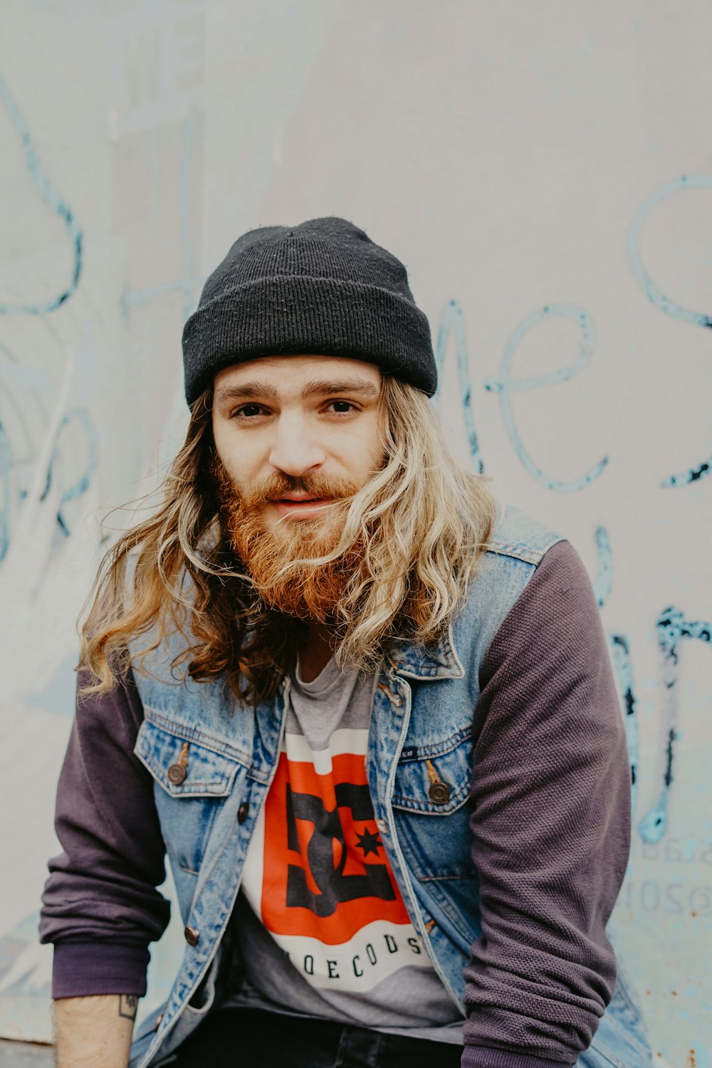 man wearing beanie hat, ddenim vest, and sweater sitting near wall