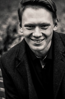 A young person with light skin and short hair is smiling while wearing a dark jacket over a shirt. The background is blurred, creating a focus on the figure's facial expression.