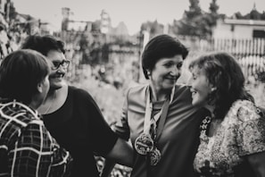 A group of four women are gathered closely together, engaged in conversation and smiling warmly. They are dressed in casual clothing and appear to be enjoying each other's company outdoors. The image is in black and white, giving it a timeless and nostalgic feel.