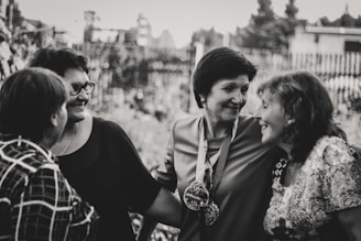 A group of four women are gathered closely together, engaged in conversation and smiling warmly. They are dressed in casual clothing and appear to be enjoying each other's company outdoors. The image is in black and white, giving it a timeless and nostalgic feel.