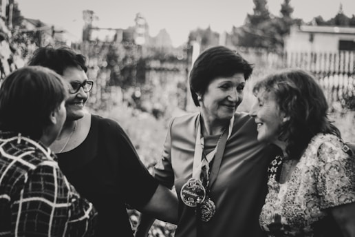 A group of four women are gathered closely together, engaged in conversation and smiling warmly. They are dressed in casual clothing and appear to be enjoying each other's company outdoors. The image is in black and white, giving it a timeless and nostalgic feel.