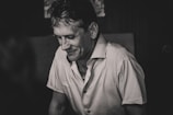 A black and white portrait of a middle-aged man looking down with a slight smile on his face. He is wearing a short-sleeved, buttoned shirt. The lighting is soft, creating gentle shadows on his features, imparting a sense of introspection or contemplation.