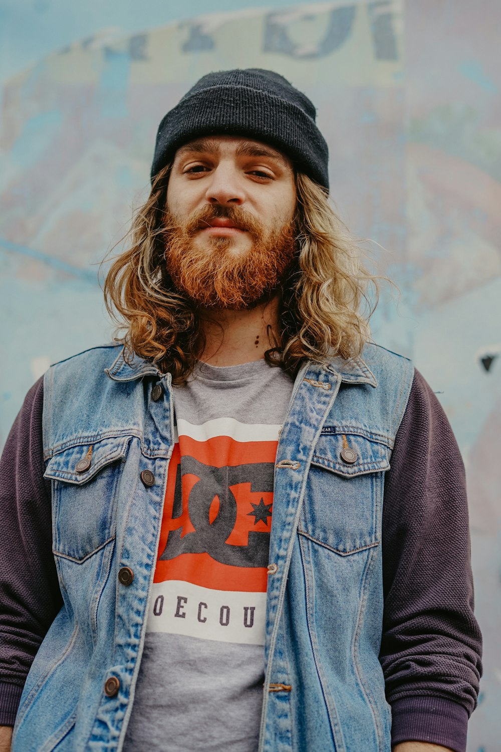 Man Wearing Beanie Hat Ddenim Vest And Sweater Sitting Near Wall
