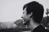A black and white profile portrait of a person with short hair, smiling and looking towards the left. In the background, there is a foggy park scene with trees and lampposts.