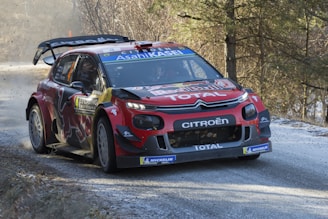 A red Citroën rally car navigates a curve on a snowy road, surrounded by trees and sparse vegetation. It features branding from Red Bull, Total, and Michelin, with visible racing decals and a rear spoiler. The car kicks up dust and snow as it speeds through the scene, suggesting a high-energy rally race setting.