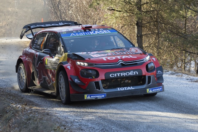 A red Citroën rally car navigates a curve on a snowy road, surrounded by trees and sparse vegetation. It features branding from Red Bull, Total, and Michelin, with visible racing decals and a rear spoiler. The car kicks up dust and snow as it speeds through the scene, suggesting a high-energy rally race setting.