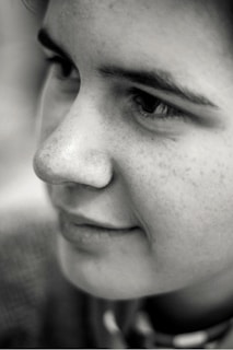 A close-up, black and white portrait of a person's face, focusing on the eyes, nose, and mouth. The lighting highlights the texture of the skin and the expression is calm and introspective.
