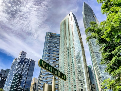 Several modern skyscrapers rise against a partly cloudy sky, with lush green trees in the foreground. A street sign reading 'Marina Blvd' is visible, adding context to the urban setting.