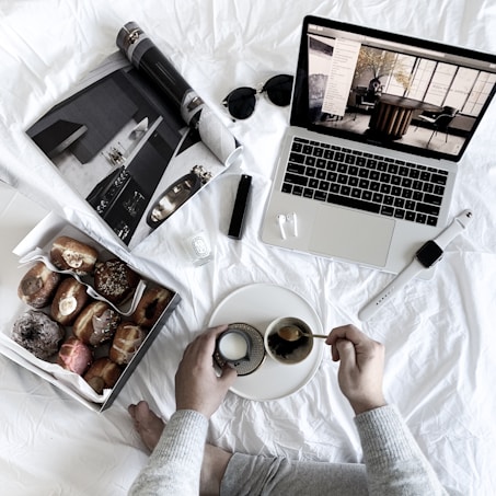 A cozy and relaxed setup featuring a person's hands holding a small cup on a plate, a box of assorted donuts, an open magazine, a laptop displaying interior design images, sunglasses, a watch, earphones, and a lighter. All items are placed on a white fabric background in a casual, comfortable manner.