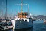 A classic white yacht docked at a marina with several smaller boats and masts visible. The yacht has a wooden cabin area and is situated under a clear blue sky. In the background, there are distant hills and other vessels.