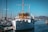 A classic white yacht docked at a marina with several smaller boats and masts visible. The yacht has a wooden cabin area and is situated under a clear blue sky. In the background, there are distant hills and other vessels.