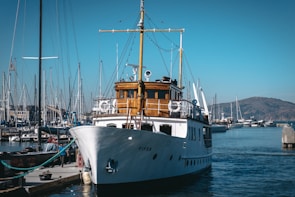 A classic white yacht docked at a marina with several smaller boats and masts visible. The yacht has a wooden cabin area and is situated under a clear blue sky. In the background, there are distant hills and other vessels.