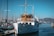 A classic white yacht docked at a marina with several smaller boats and masts visible. The yacht has a wooden cabin area and is situated under a clear blue sky. In the background, there are distant hills and other vessels.