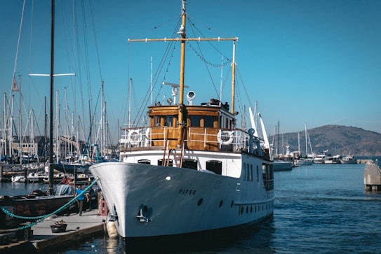 A classic white yacht docked at a marina with several smaller boats and masts visible. The yacht has a wooden cabin area and is situated under a clear blue sky. In the background, there are distant hills and other vessels.
