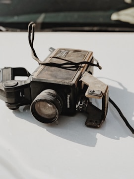 A vintage Panasonic video camera with a worn and dusty appearance is placed on a smooth, light-colored surface. The camera features a prominent lens and several buttons and controls, alongside old straps.