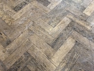 A close-up view of a wooden parquet floor with a herringbone pattern. The wood has a light brown color with visible grain and a slightly worn appearance.