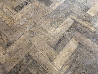 A close-up view of a wooden parquet floor with a herringbone pattern. The wood has a light brown color with visible grain and a slightly worn appearance.