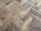 A close-up view of a wooden parquet floor with a herringbone pattern. The wood has a light brown color with visible grain and a slightly worn appearance.