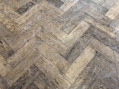 A close-up view of a wooden parquet floor with a herringbone pattern. The wood has a light brown color with visible grain and a slightly worn appearance.