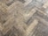 A close-up view of a wooden parquet floor with a herringbone pattern. The wood has a light brown color with visible grain and a slightly worn appearance.