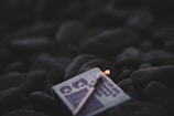 A close-up of a matchbox resting on dark, smooth stones with a single lit matchstick placed on top. The surrounding area appears dimly lit, emphasizing the soft glow of the match flame.