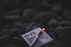 A close-up of a matchbox resting on dark, smooth stones with a single lit matchstick placed on top. The surrounding area appears dimly lit, emphasizing the soft glow of the match flame.