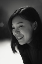 A black and white portrait of a young person smiling, with shoulder-length dark hair. The lighting is soft, highlighting the features gently.