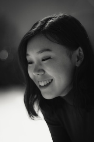 A black and white portrait of a young person smiling, with shoulder-length dark hair. The lighting is soft, highlighting the features gently.