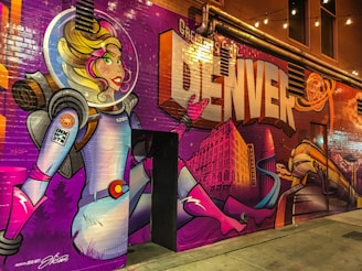 A vibrant mural featuring a female astronaut with colorful hair in a spacesuit, against a background of large lettering spelling out 'DENVER'. The artwork includes elements of a cityscape, with a prominent building and a train. The scene is illuminated by string lights above, adding to the lively ambience.