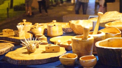 A variety of hand-crafted wooden kitchenware items are displayed on a table, including bowls, mortars and pestles, toothpick holders, and other small containers. These items are made from light-colored wood, and the natural grain patterns are visible. The setup is outdoors, likely at a market or fair, with a dim evening lighting that casts warm shadows.