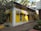 A yellow building with multiple air conditioning units attached to its walls is situated along a narrow walkway. There are trees and plants surrounding the building, with palm trees visible in the background.