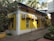 A yellow building with multiple air conditioning units attached to its walls is situated along a narrow walkway. There are trees and plants surrounding the building, with palm trees visible in the background.