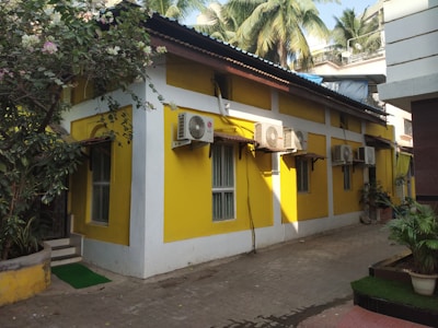 A yellow building with multiple air conditioning units attached to its walls is situated along a narrow walkway. There are trees and plants surrounding the building, with palm trees visible in the background.