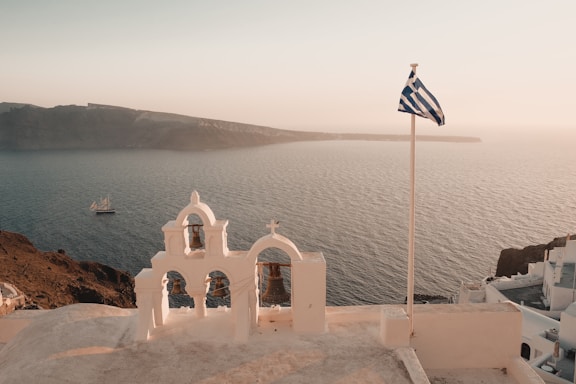 An iconic Greek church with whitewashed structures and blue domes overlooks the sea. A Greek flag flutters in the breeze, near intricate bells on top of the church. The backdrop features a serene ocean view with a distant island and a sailing ship.