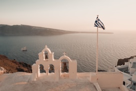 An iconic Greek church with whitewashed structures and blue domes overlooks the sea. A Greek flag flutters in the breeze, near intricate bells on top of the church. The backdrop features a serene ocean view with a distant island and a sailing ship.
