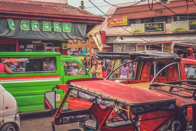 A bustling street market scene with colorful jeepneys and tricycles occupying the road. People are walking around, some stopping at various stalls and shops. The market is vibrant and lively, with signs and advertisements visible on the buildings.