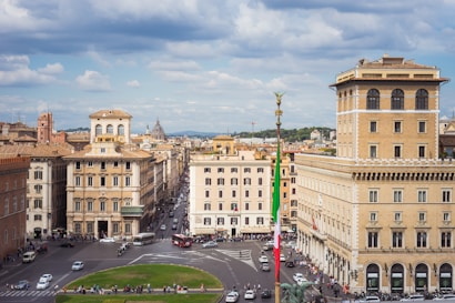 A bustling urban scene features historic buildings surrounding a central square. The architecture is characterized by classical style facades with arched windows and decorative details. A tall flagpole with the Italian flag stands prominently in the foreground. Streets are filled with cars and pedestrians, and the scene conveys a sense of vibrant city life.