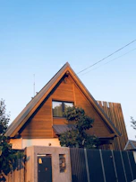 A wooden A-frame house with a steep triangular roof, featuring a large window in the upper portion. The house is surrounded by various plants and trees, and there is a modern fence with a large gate in the foreground. The sky is clear and blue.