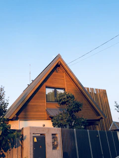 A wooden A-frame house with a steep triangular roof, featuring a large window in the upper portion. The house is surrounded by various plants and trees, and there is a modern fence with a large gate in the foreground. The sky is clear and blue.