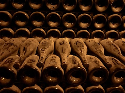 Rows of dusty wine bottles are stacked horizontally in a dimly lit cellar. The bottles are arranged neck-out, with various markings and labels visible on each. The atmosphere is one of age and preservation, with a warm, sepia-toned lighting casting shadows and highlighting the contours of the bottles.