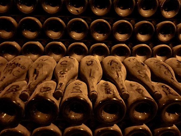 Rows of dusty wine bottles are stacked horizontally in a dimly lit cellar. The bottles are arranged neck-out, with various markings and labels visible on each. The atmosphere is one of age and preservation, with a warm, sepia-toned lighting casting shadows and highlighting the contours of the bottles.