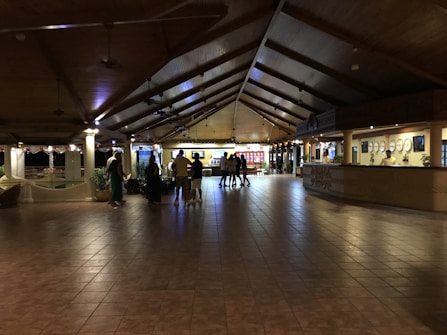 A spacious hotel lobby with a high wooden ceiling and tiled floor. Several people are gathered near a counter, possibly checking in or out. The lighting is warm and dim, creating a cozy atmosphere. There are decorative elements such as plants and wall art. The reception desk is staffed and well-lit, and there is a seating area visible in the background.